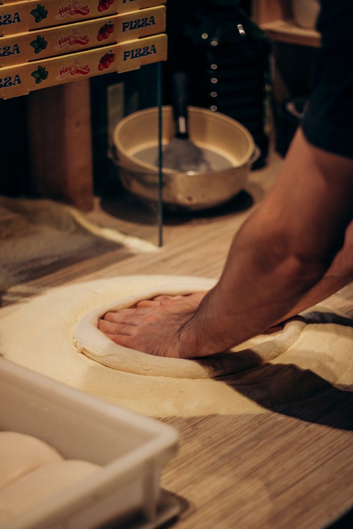 A person kneading pizza dough on a wooden countertop with ingredients nearby, in a warm kitchen environment.